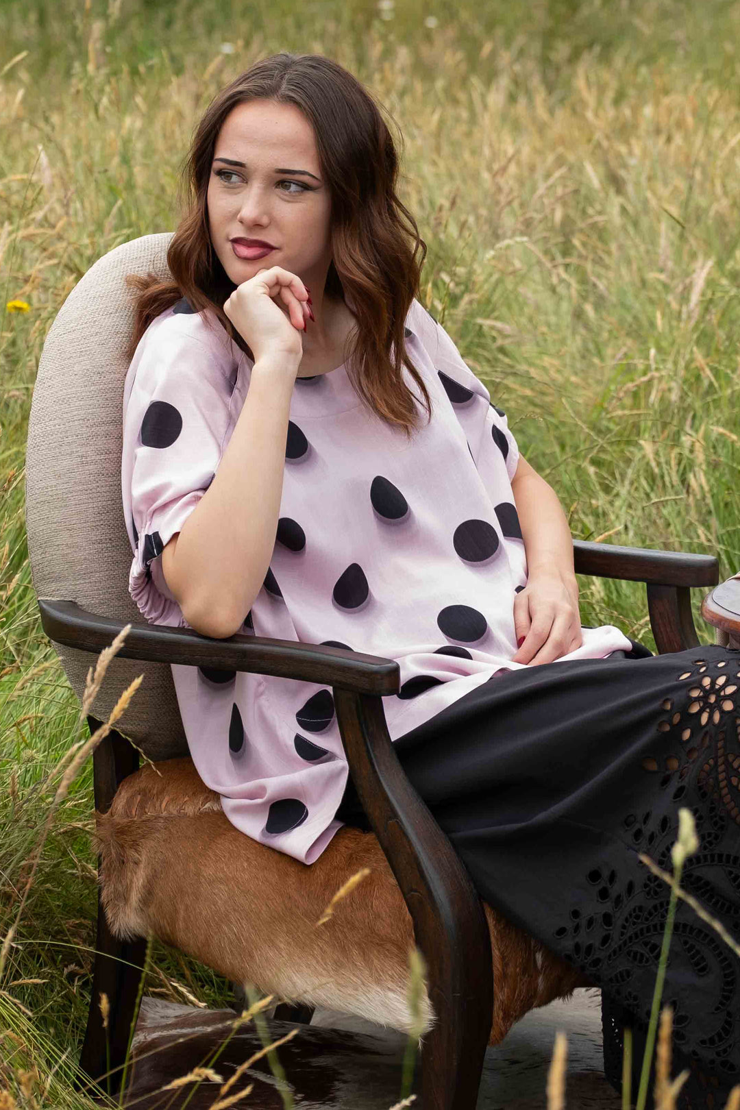 A woman sits in a rustic outdoor chair in a grassy field, wearing a light pink top with large black polka dots and a long black skirt. A small wooden table with a book is beside her.