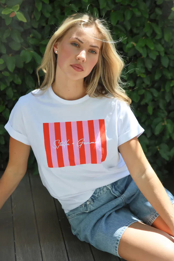 A blonde woman sitting on a wooden deck in front of a green hedge, wearing a white t-shirt with a red and pink striped graphic and denim shorts.