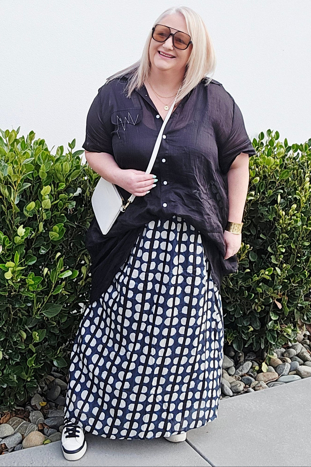A woman wearing a black short-sleeve button-up shirt and a navy and white patterned maxi skirt, standing outdoors on a concrete path next to green bushes against a white wall.