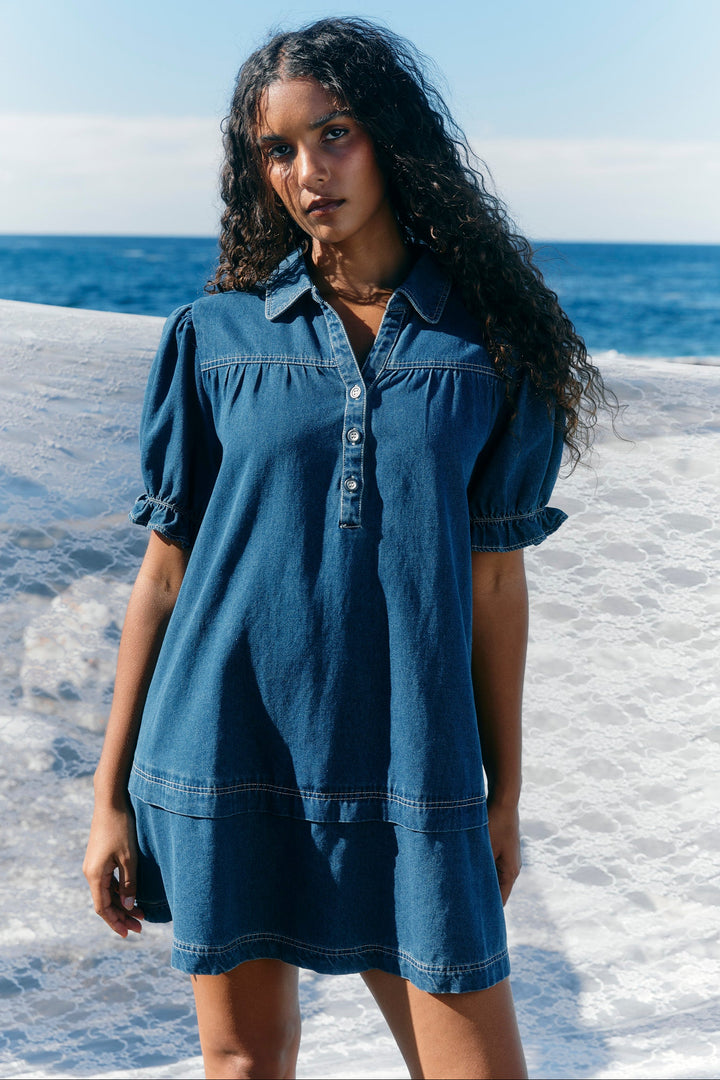 A woman with long, curly dark hair stands outdoors by the ocean wearing a blue denim shirt dress. The dress has short puff sleeves, a buttoned collar, and a tiered skirt.