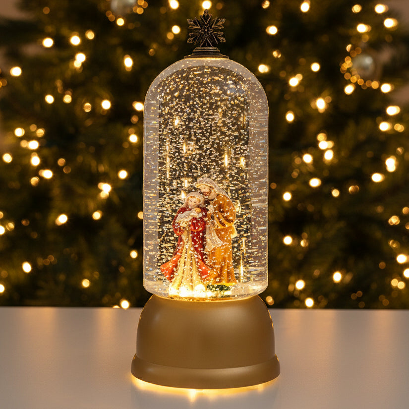 A golden snow globe figurine featuring Santa Claus and Mrs. Claus under a clear glass dome with glittering lights, set on a light gray surface with a blurred snowy pine branch in the background.
