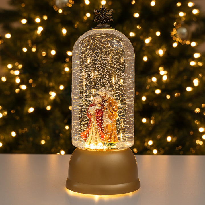 A golden snow globe figurine featuring Santa Claus and Mrs. Claus under a clear glass dome with glittering lights, set on a light gray surface with a blurred snowy pine branch in the background.
