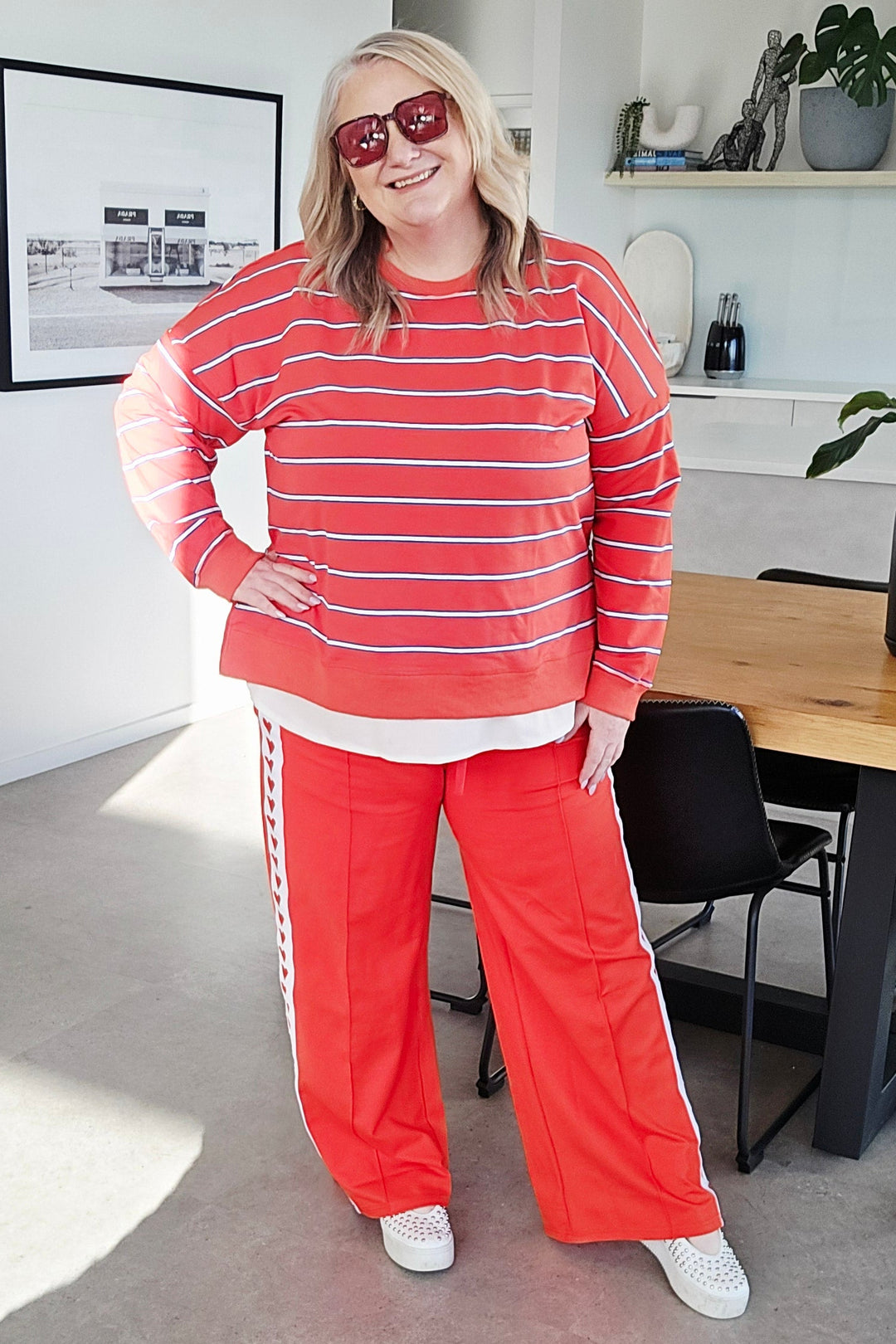 A woman wearing a matching red and white striped long-sleeve top and red pants with white side stripes, standing in an indoor room with a gray carpet and office furniture in the background.