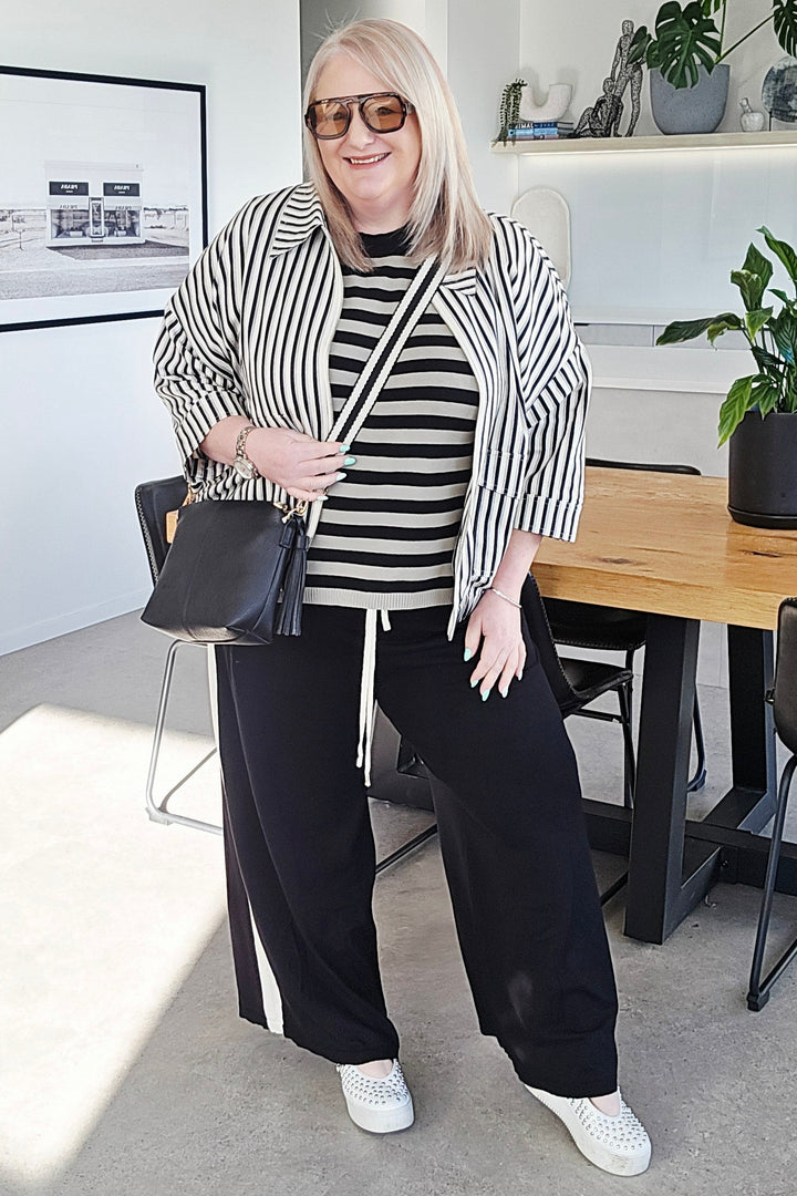 A woman stands in a modern indoor room wearing a black and white striped blazer over a matching striped top, black wide-leg pants with white side stripes, white sneakers, and holding a black handbag.