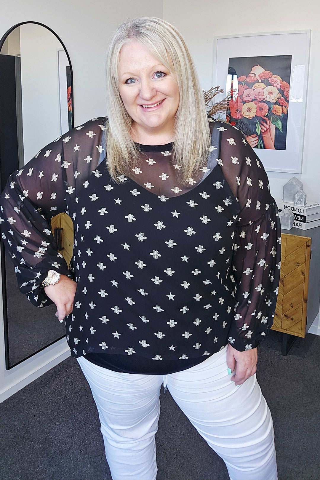 Person wearing a black patterned top and white pants in a room with a mirror and decor.