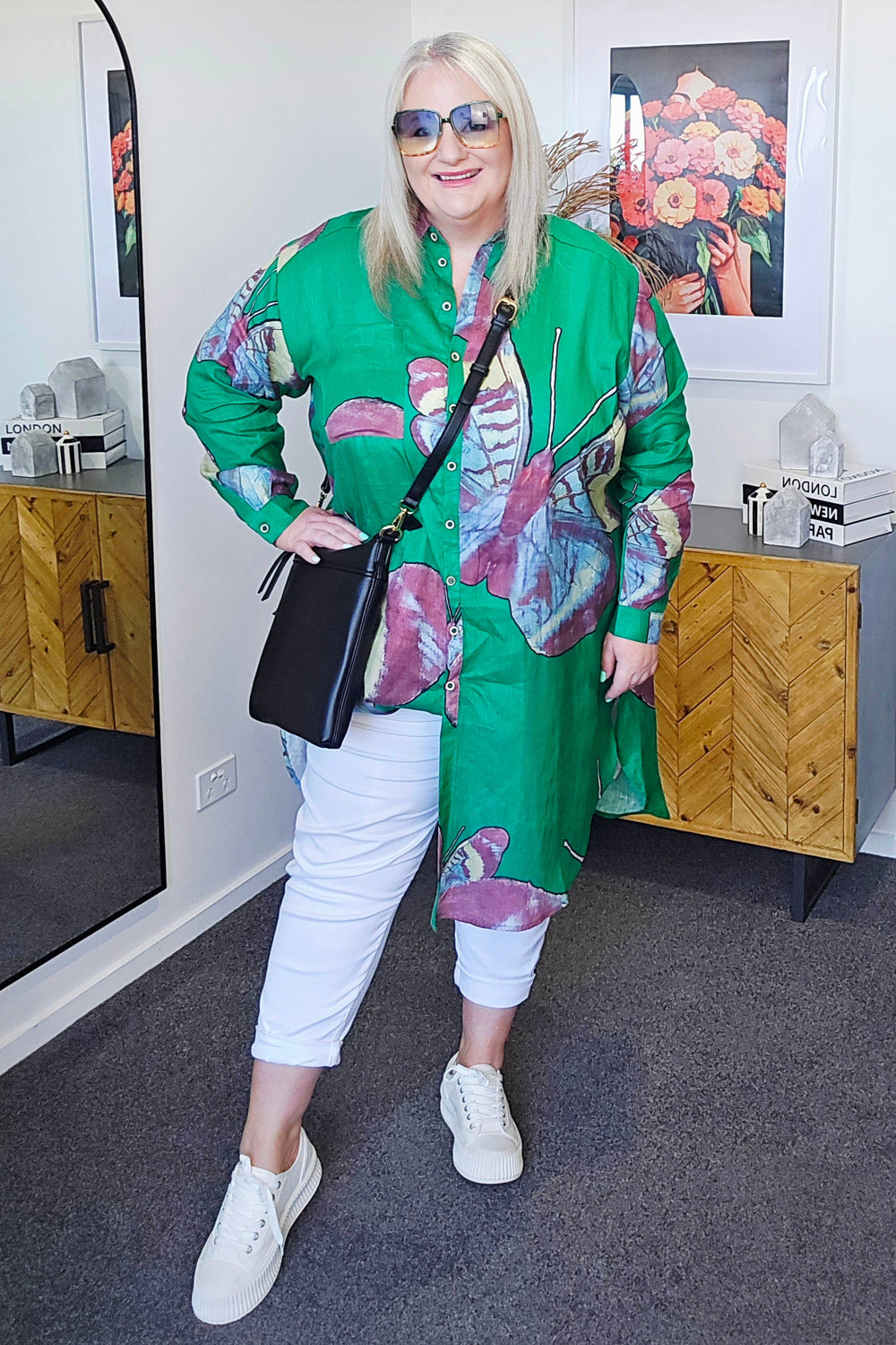 Person wearing a colorful outfit with a green jacket and white pants, standing in a room with wooden furniture and a mirror.