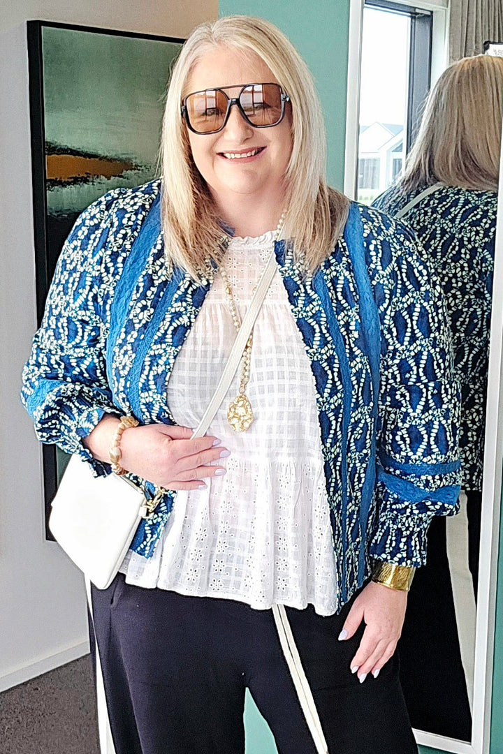 Woman in a stylish outfit with a blue patterned jacket and black pants, standing in a room with a mirror.