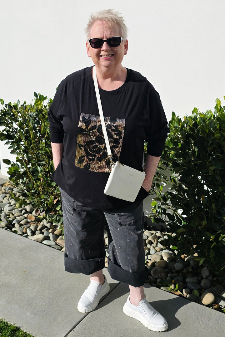 A woman wearing a black graphic t-shirt, dark gray cropped pants, and white sneakers stands outdoors on a concrete walkway next to a white building and green bushes, carrying a small white shoulder bag.