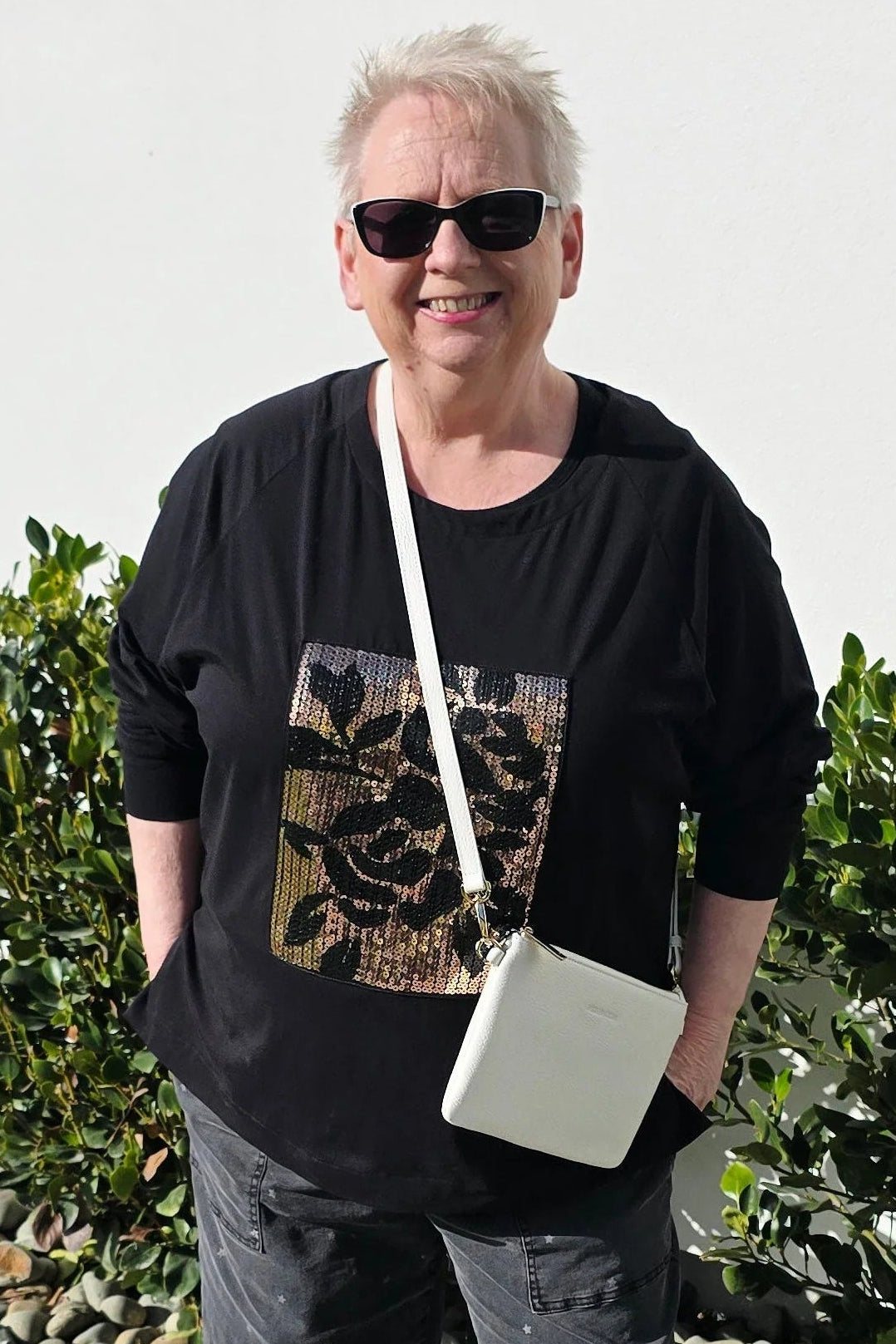 A woman wearing a black graphic t-shirt, dark cropped pants, white slip-on shoes, and a white crossbody bag, standing outdoors on a concrete path with green bushes behind her.