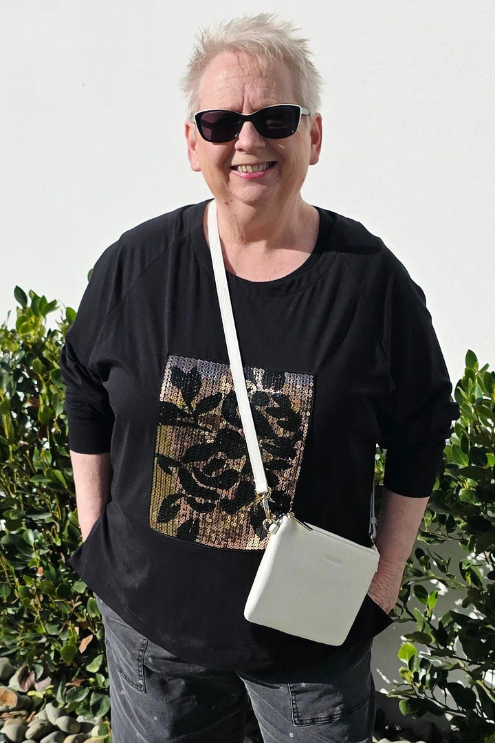 A woman wearing a black graphic t-shirt, dark cropped pants, white slip-on shoes, and a white crossbody bag, standing outdoors on a concrete path with green bushes behind her.