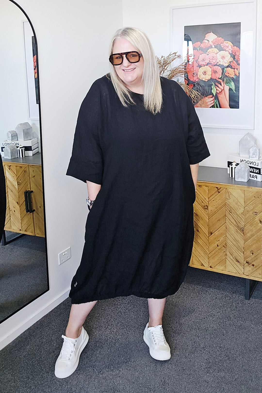 Person wearing a plus size black dress standing in a room with a mirror reflecting the interior.