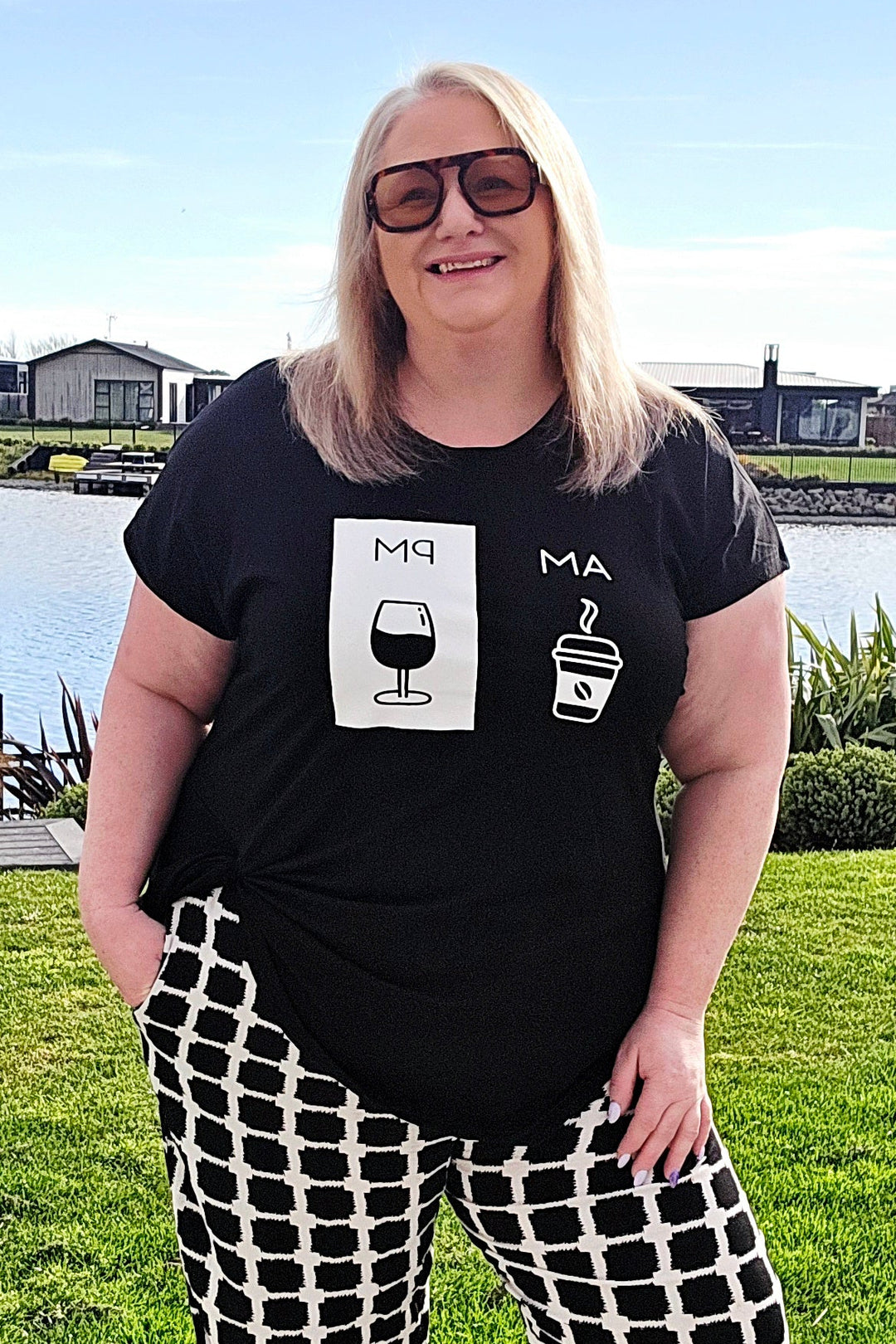 A woman wearing a black graphic t-shirt and black-and-white checkered wide-leg pants stands outdoors on a concrete walkway with a grassy lawn, water, and houses in the background.