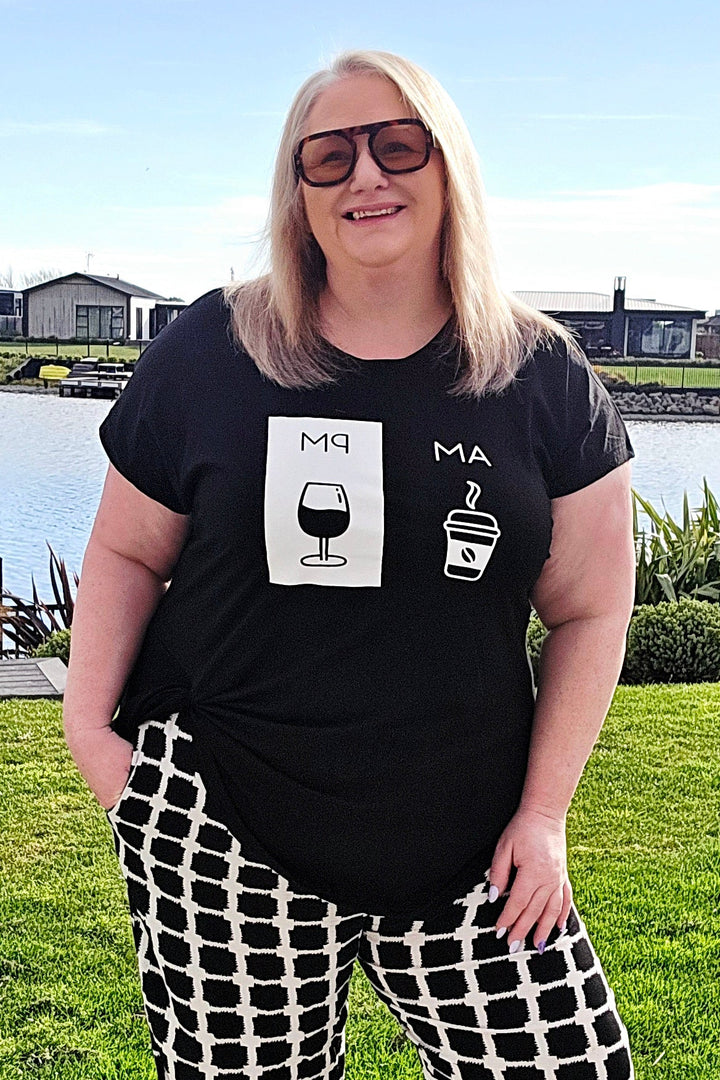 A woman wearing a black graphic t-shirt and black-and-white checkered wide-leg pants stands outdoors on a concrete walkway with a grassy lawn, water, and houses in the background.