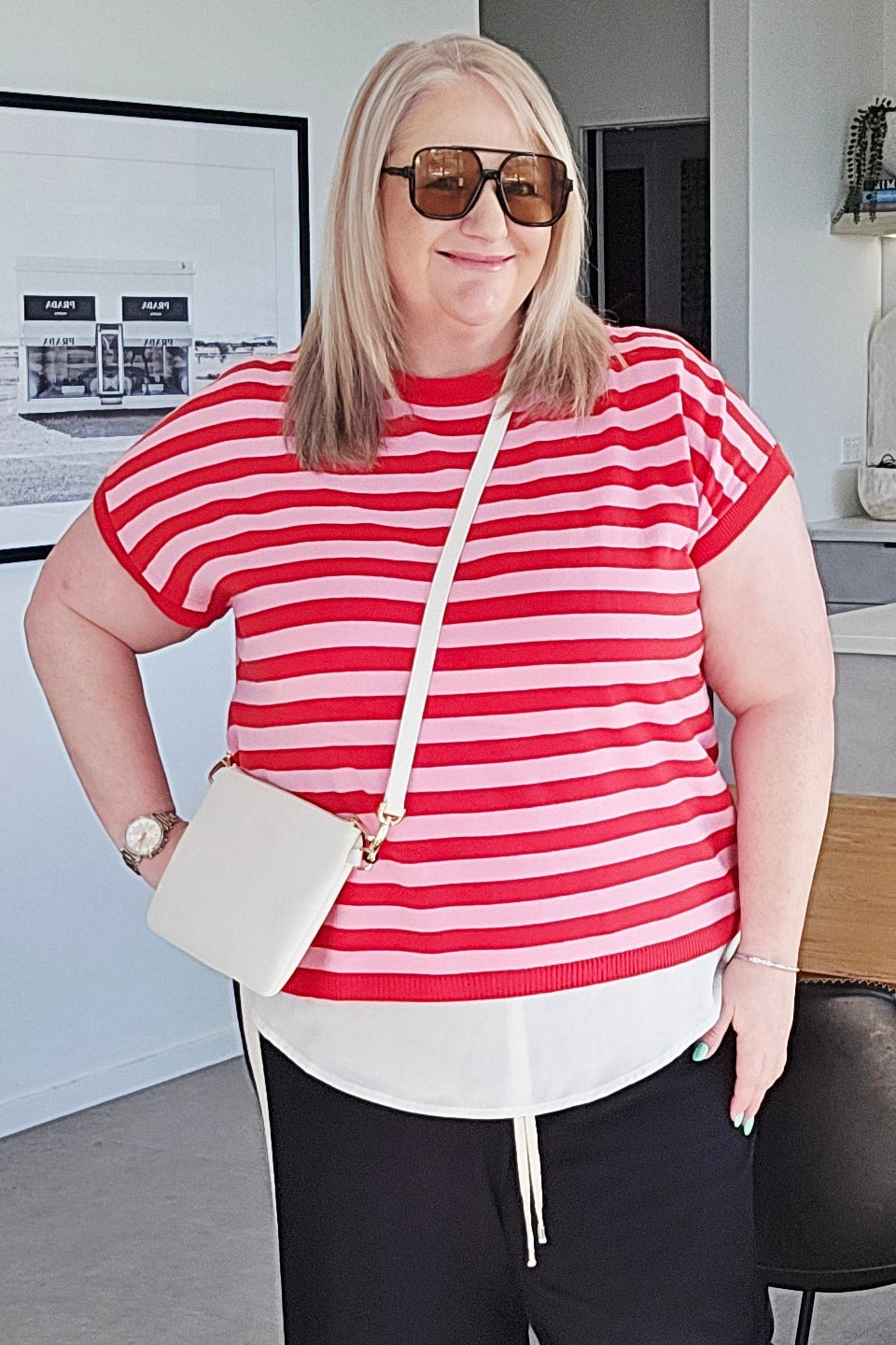 A woman wearing a red and white striped short-sleeve top, black wide-leg pants, and a white crossbody bag stands in a modern indoor setting with a dining table and plants in the background.