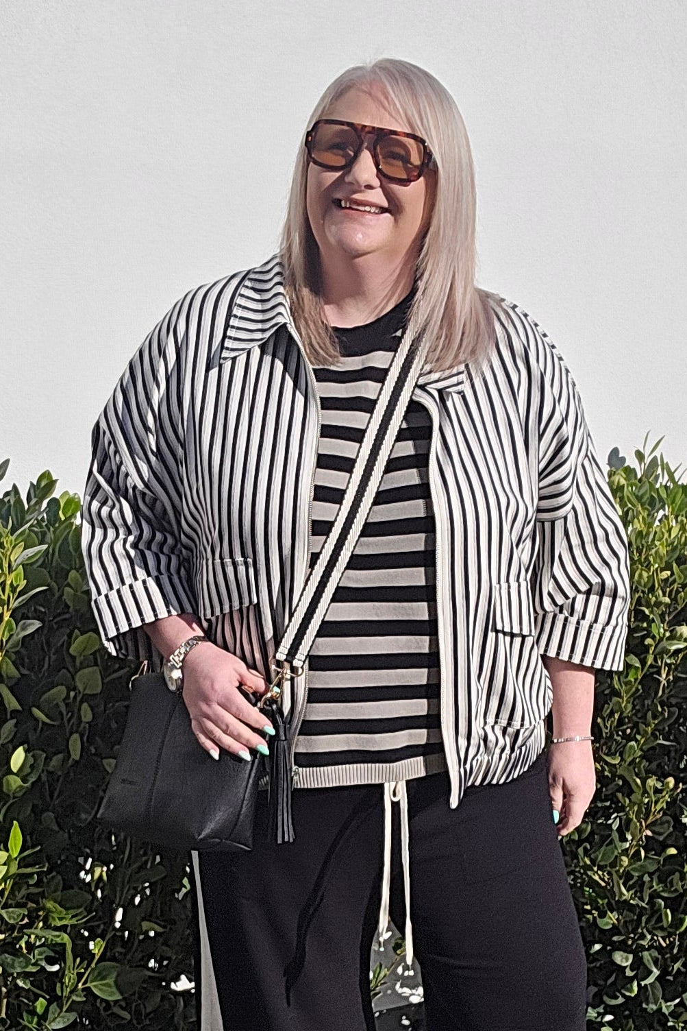 A woman stands outdoors on a concrete walkway in front of a hedge, wearing a black and white vertically striped button-up shirt, a matching striped t-shirt, black wide-leg pants with white side stripes, and white sneakers.