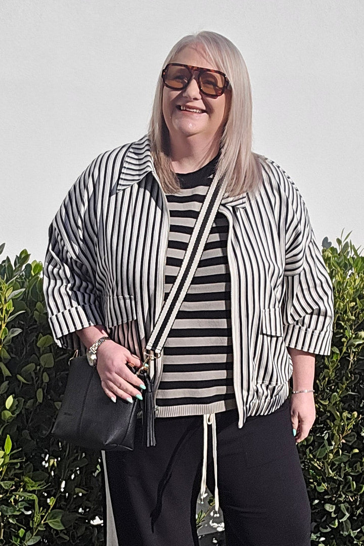 A woman stands outdoors on a concrete walkway in front of a hedge, wearing a black and white vertically striped button-up shirt, a matching striped t-shirt, black wide-leg pants with white side stripes, and white sneakers.