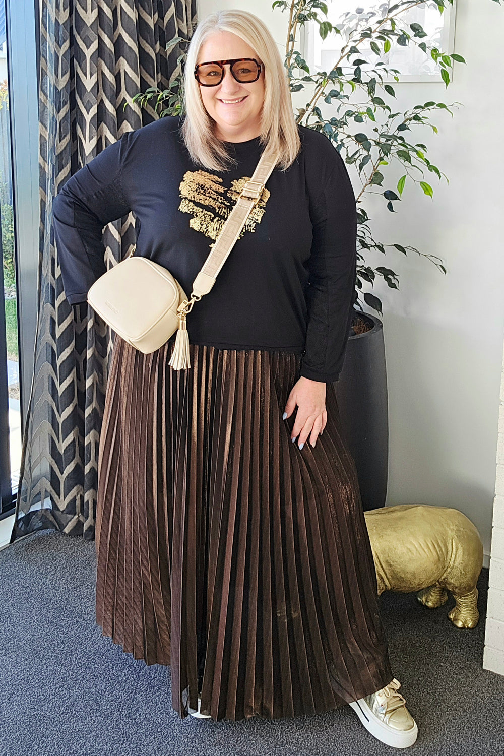 Woman wearing a black top and brown pleated skirt with sunglasses indoors.