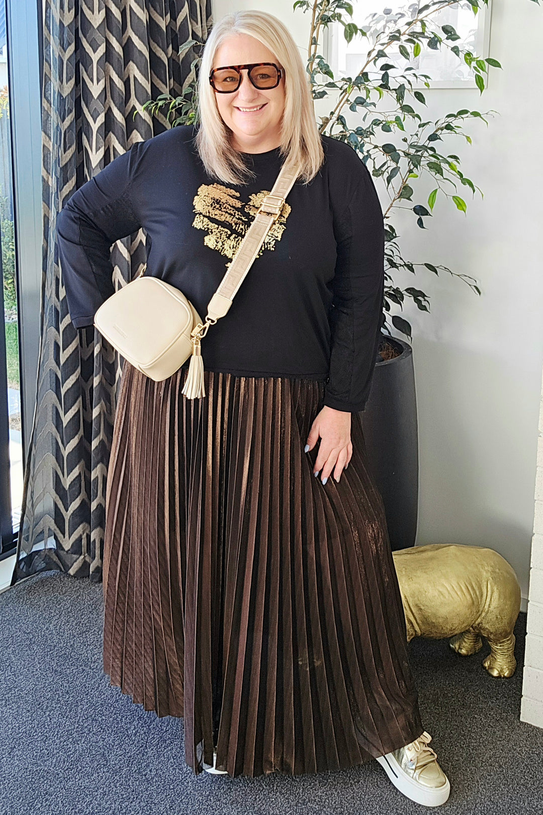 Woman wearing a black top and brown pleated skirt with sunglasses indoors.