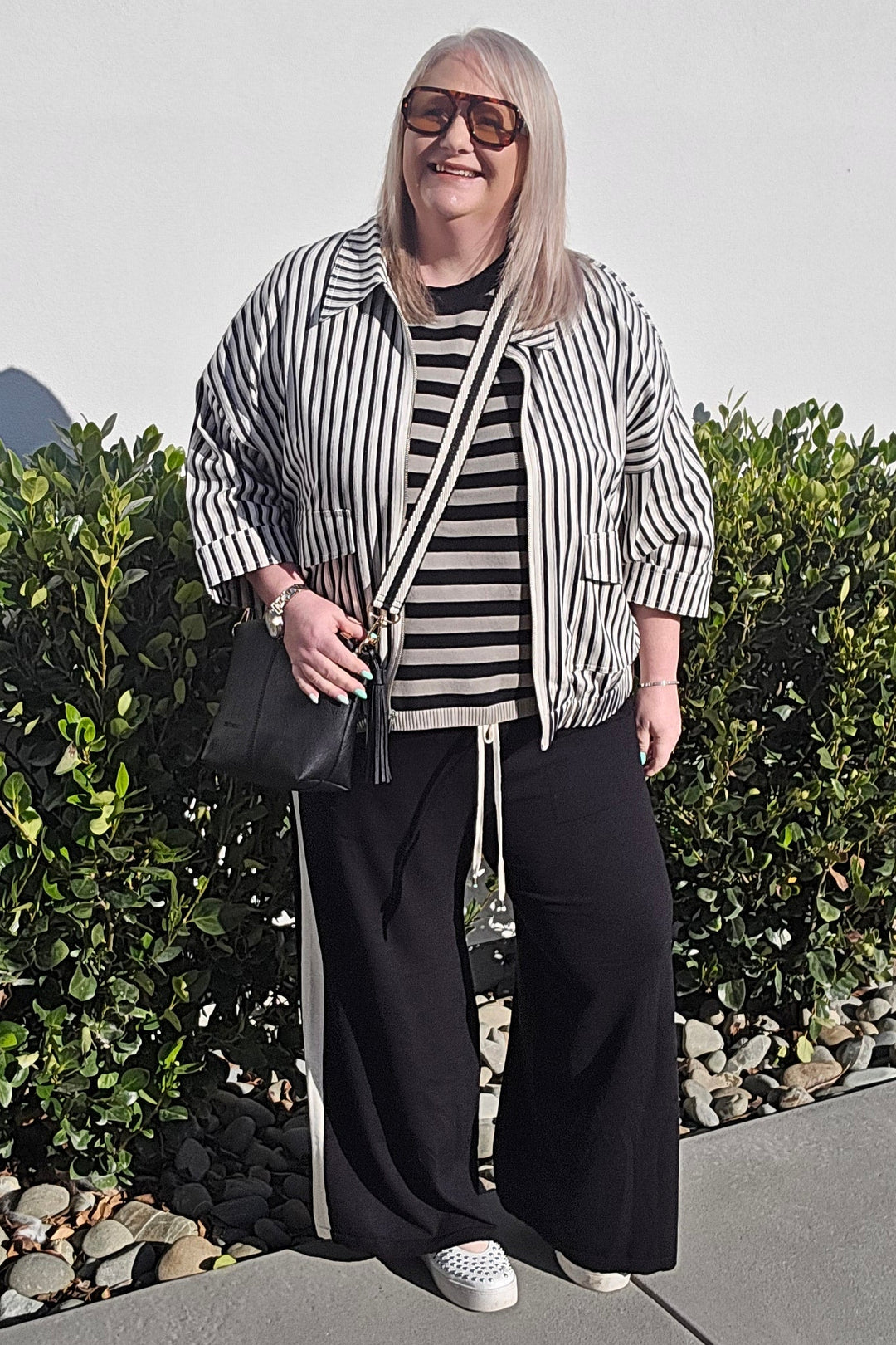 A woman stands outdoors on a concrete walkway in front of a hedge, wearing a black and white vertically striped button-up shirt, a matching striped t-shirt, black wide-leg pants with white side stripes, and white sneakers.