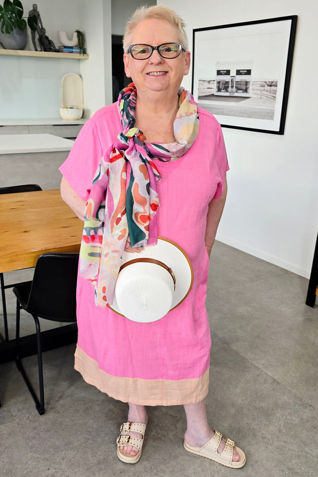 A woman with short blonde hair and glasses stands in a modern room wearing a bright pink dress, a colorful patterned scarf, and holding a white sun hat with a tan band.