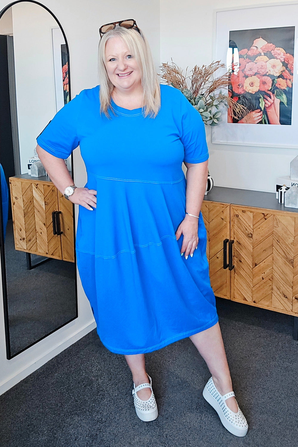 Woman in a plus size blue dress standing in a room with a mirror and wooden cabinet.