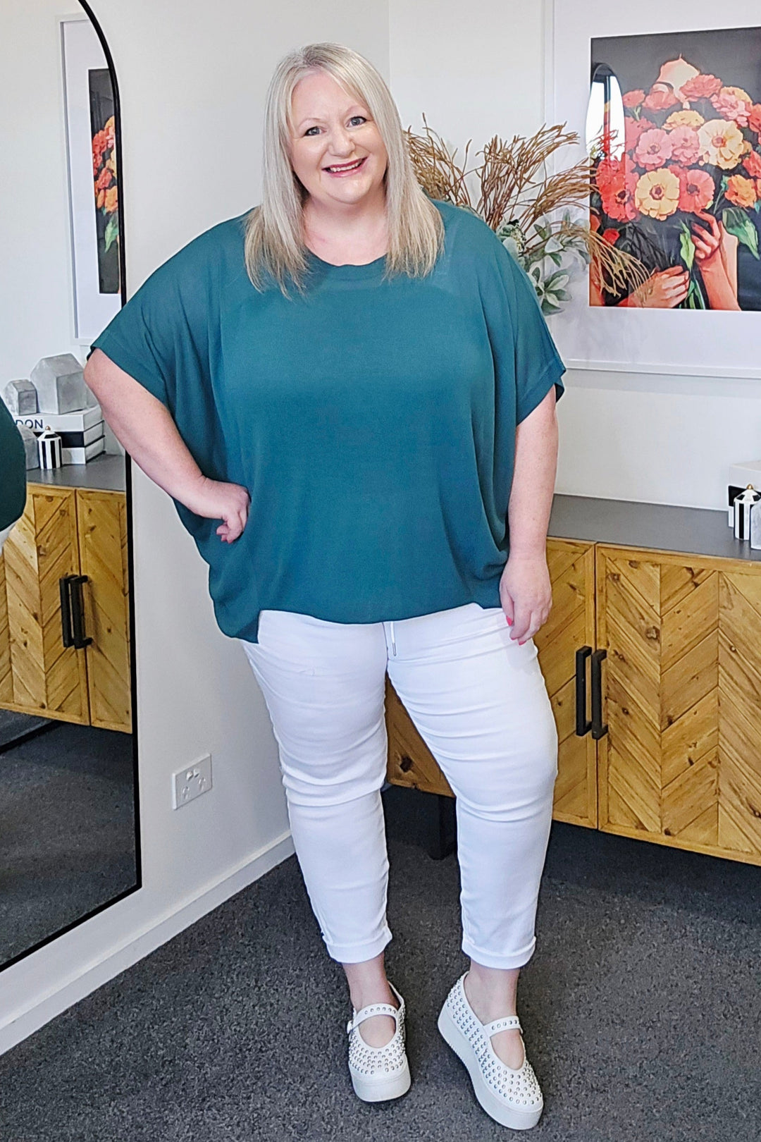 Woman wearing a teal top and white pants standing in a room with wooden furniture and floral decorations.