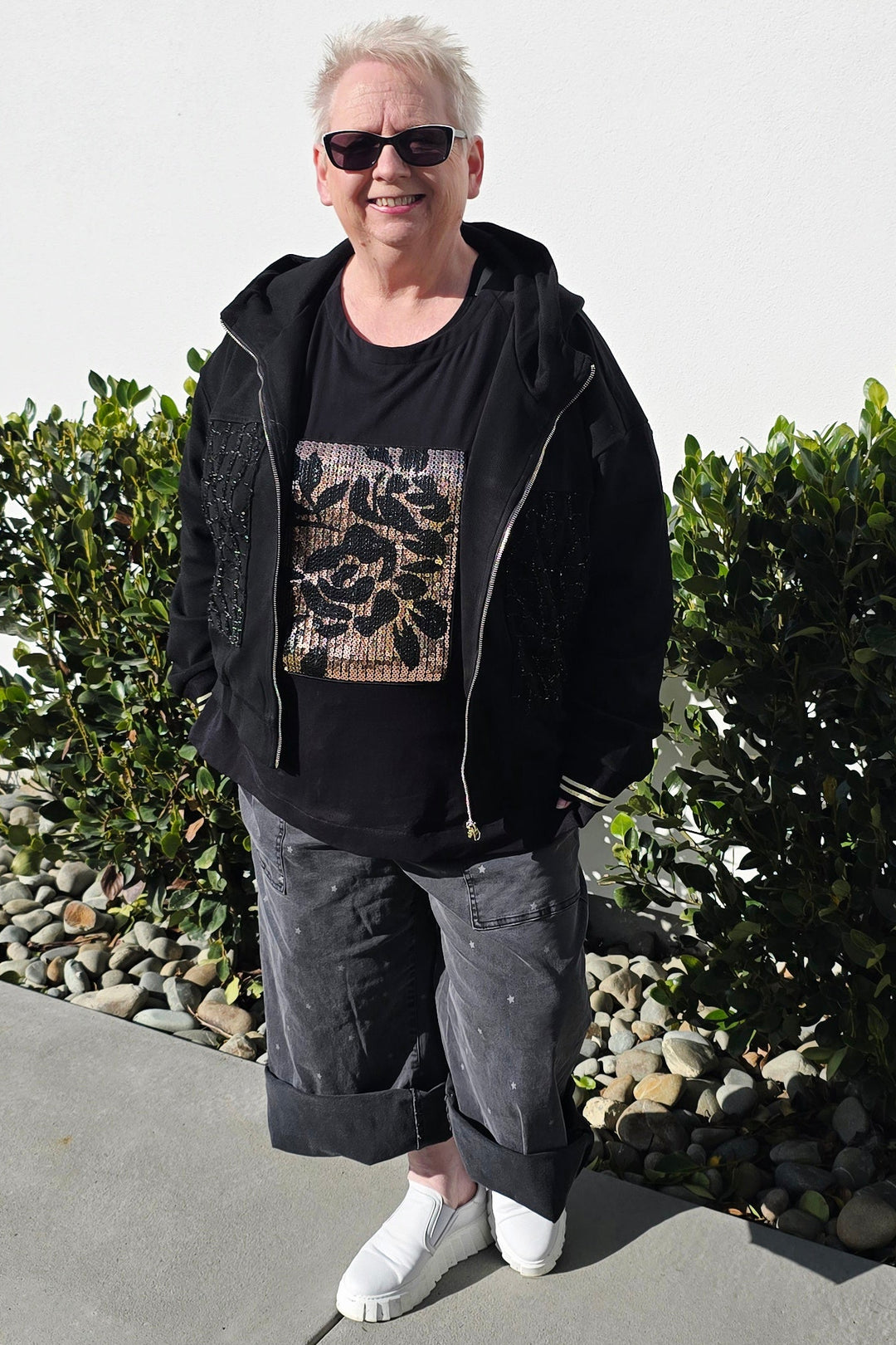 A person standing outdoors on a concrete walkway in front of a white building, wearing a black zip-up jacket over a black graphic T-shirt, dark gray rolled-up pants, and white sneakers.