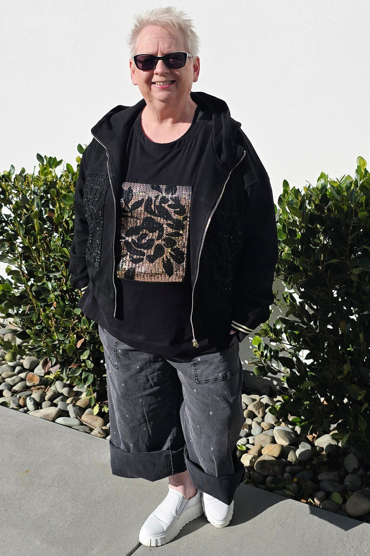 A person standing outdoors on a concrete walkway in front of a white building, wearing a black zip-up jacket over a black graphic T-shirt, dark gray rolled-up pants, and white sneakers.