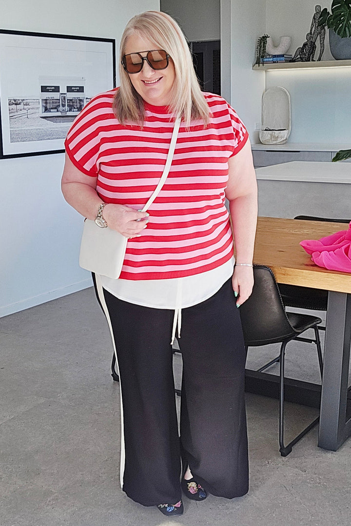 A woman wearing a red and white striped short-sleeve top, black wide-leg pants with white side stripes, and carrying a white shoulder bag, standing in a modern indoor setting.