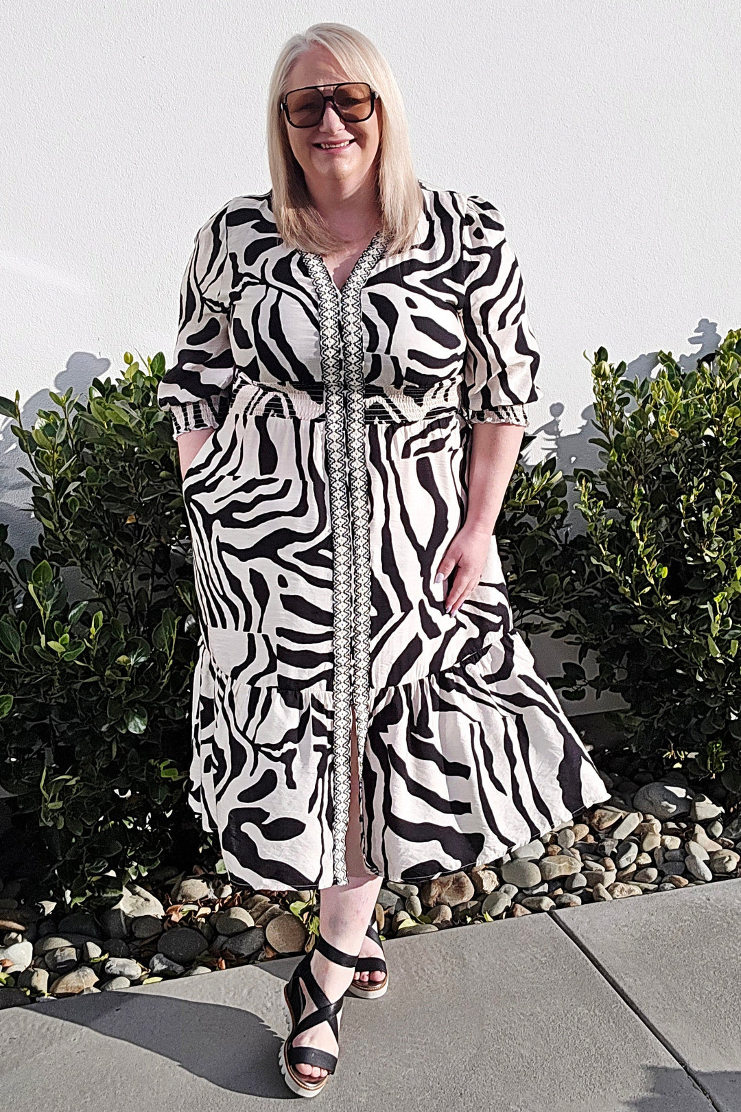 A woman wearing a black and white zebra-print midi dress with a button-up front and ruffled hem, standing outdoors on a concrete patio with green shrubs in the background.