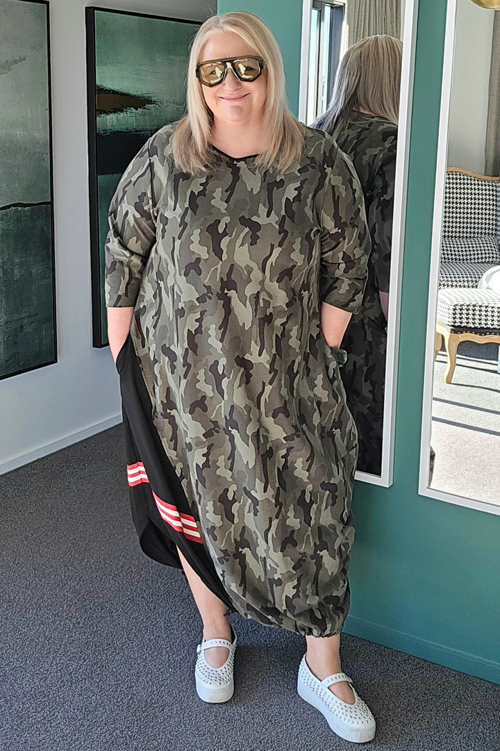 Woman wearing a camouflage dress standing in a room with a mirror reflecting her image.