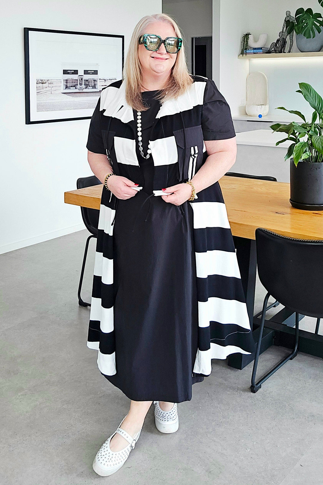 A woman wearing a black and white horizontally striped midi dress with a white collar and white shoes, standing in a modern dining room setting.