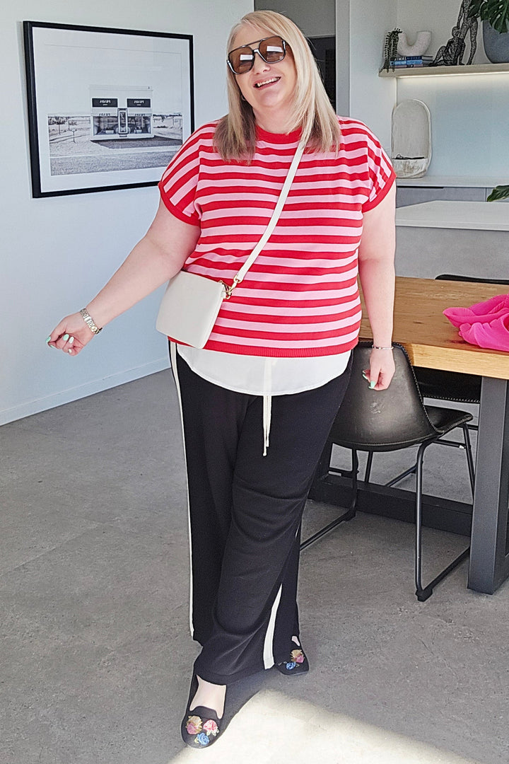 A woman wearing a red and white striped t-shirt, black sweatpants, and a white crossbody bag stands in a modern room with a dining table and plants.