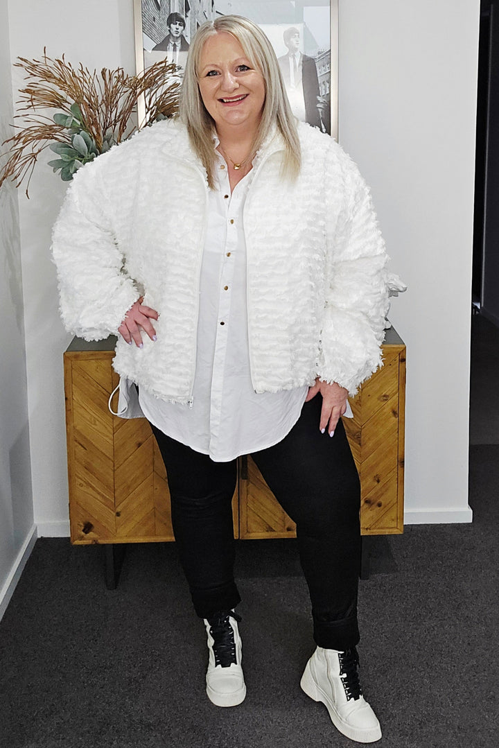 Woman wearing a white coat in an indoor setting with a plant and framed picture on the wall.