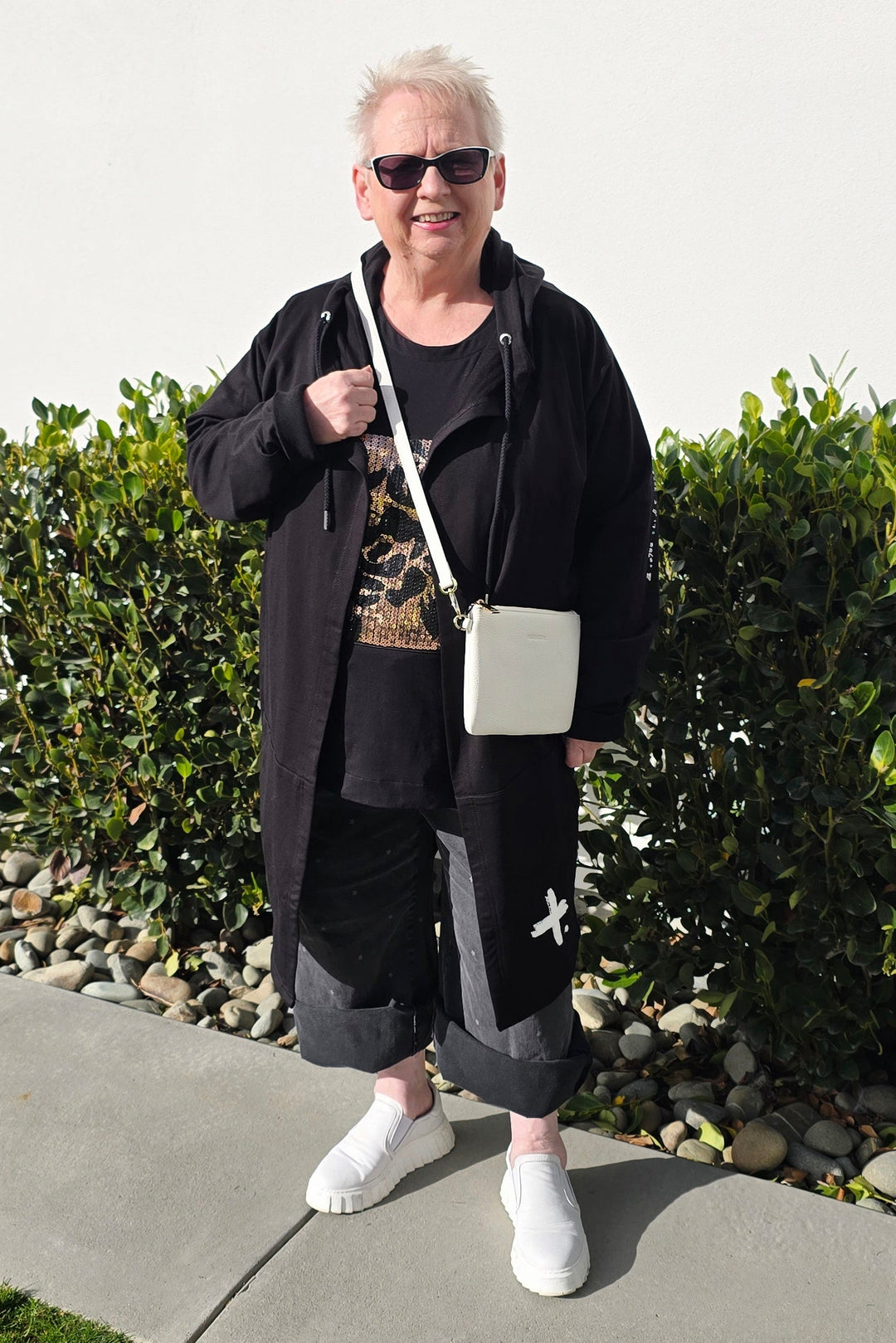 An older woman stands outdoors on a concrete walkway in front of a white building, wearing a long black coat over a black top and black pants, with white sneakers and a small white shoulder bag.