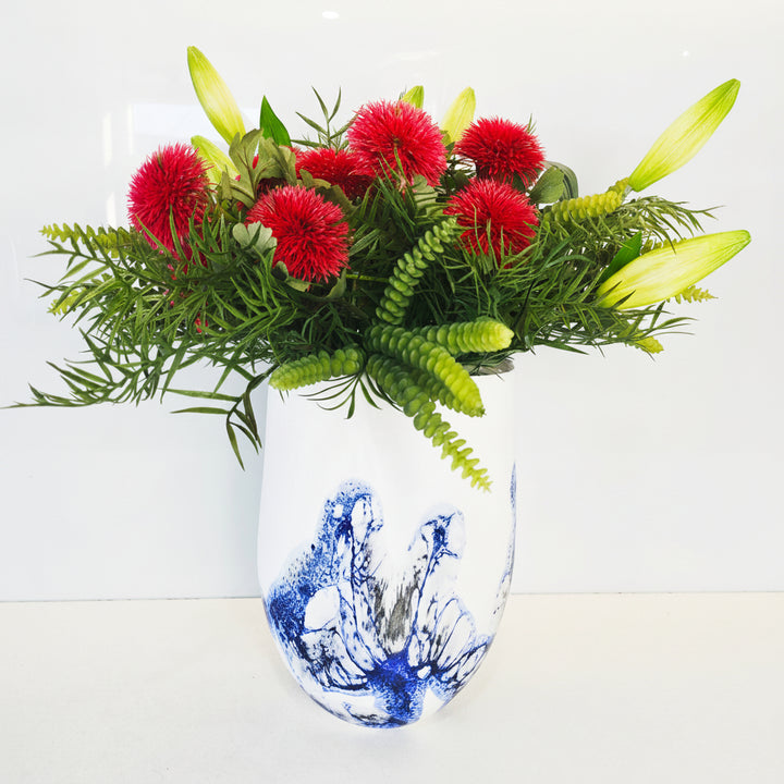 A ceramic vase with a blue and white hand-painted style design, filled with a vibrant floral arrangement featuring bright red pom-pom flowers, green ferns, and yellow lily buds, set against a plain light background.