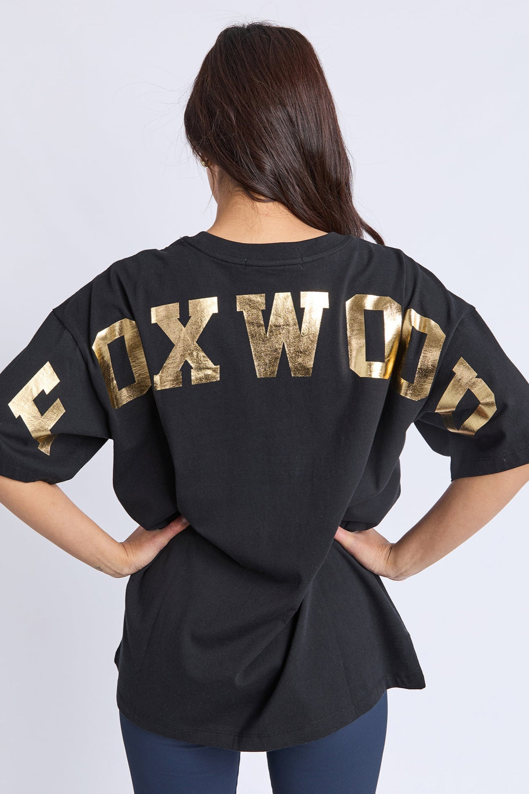 Person wearing a black t-shirt with 'FOXWOOD' printed in gold on the back against a white background