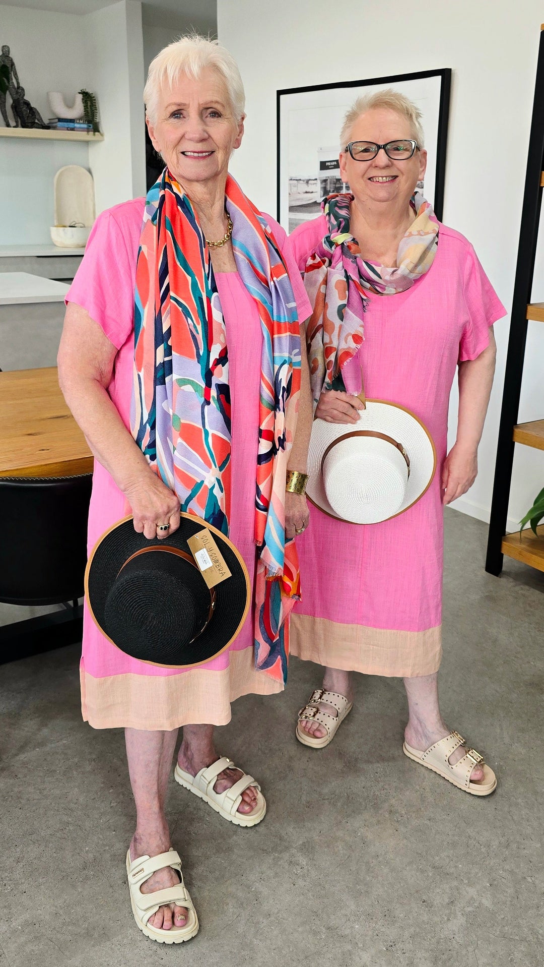 Two women standing indoors wearing matching bright pink dresses with beige hems and holding hats. One woman has a colorful abstract scarf, and the other wears a neutral scarf.