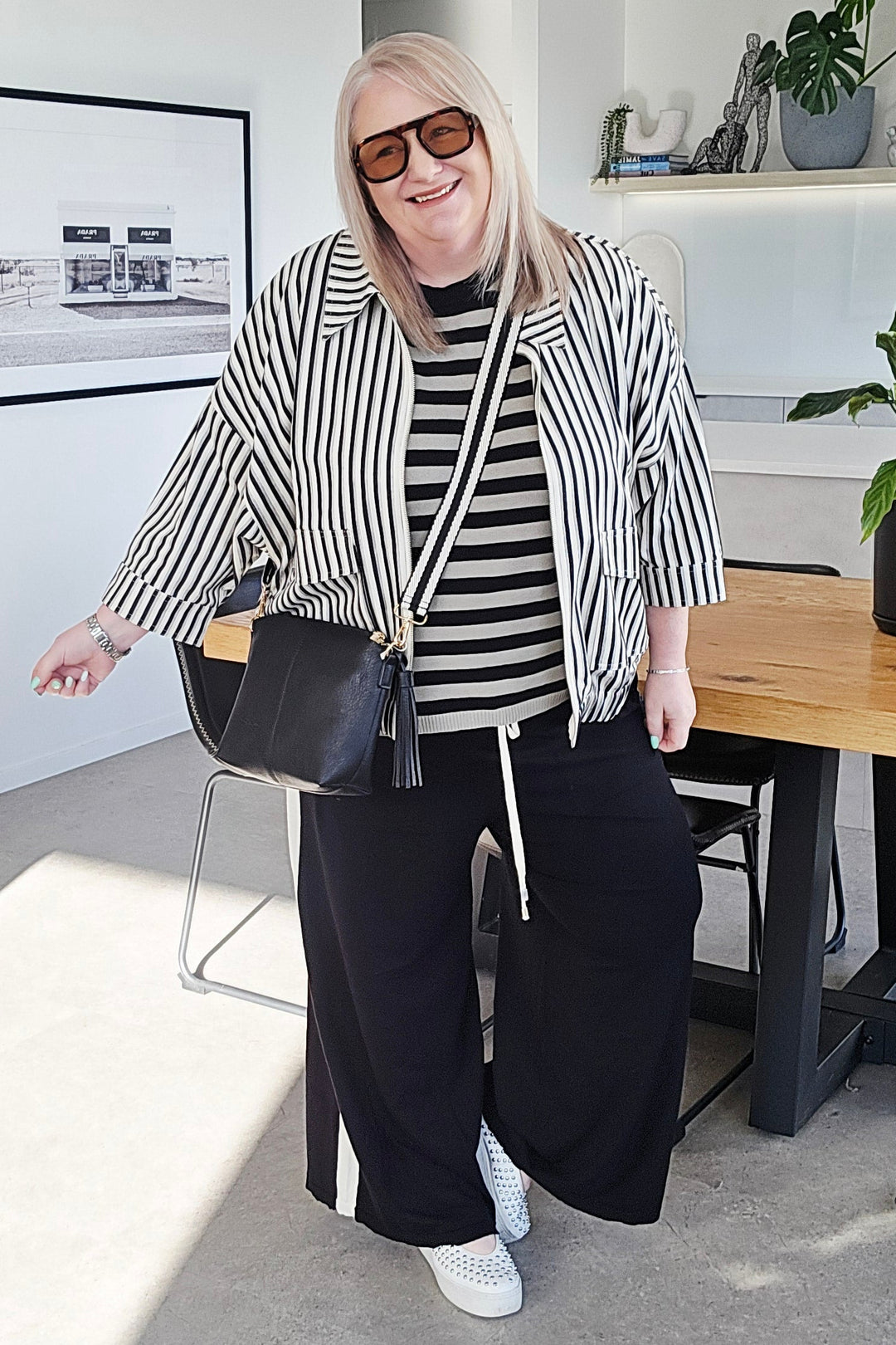 A woman wearing a black and white striped cardigan, black and white striped t-shirt, black wide-leg pants, and white sneakers stands in a modern room, holding a black shoulder bag.