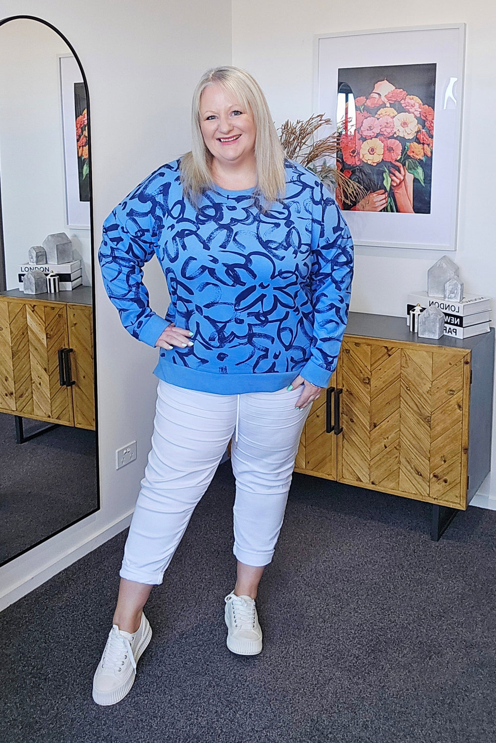 Woman wearing a blue patterned sweater and white pants standing in a room with wooden furniture and framed pictures.