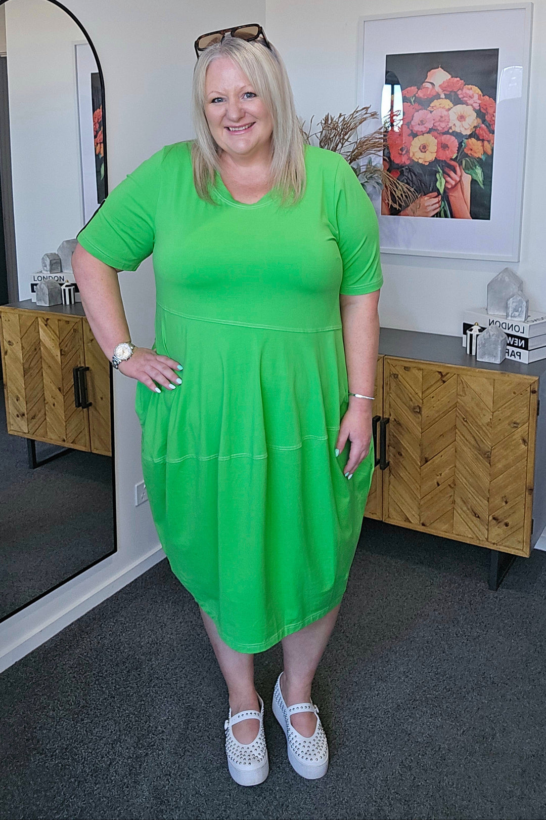 Woman in a plus size green dress standing in a room with a mirror and decorative items.