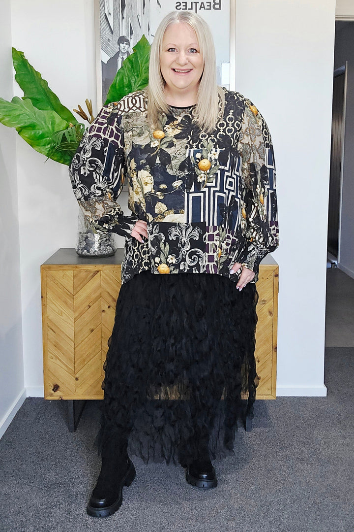 Woman in a patterned top and black skirt standing in a room with a plant and wooden cabinet.