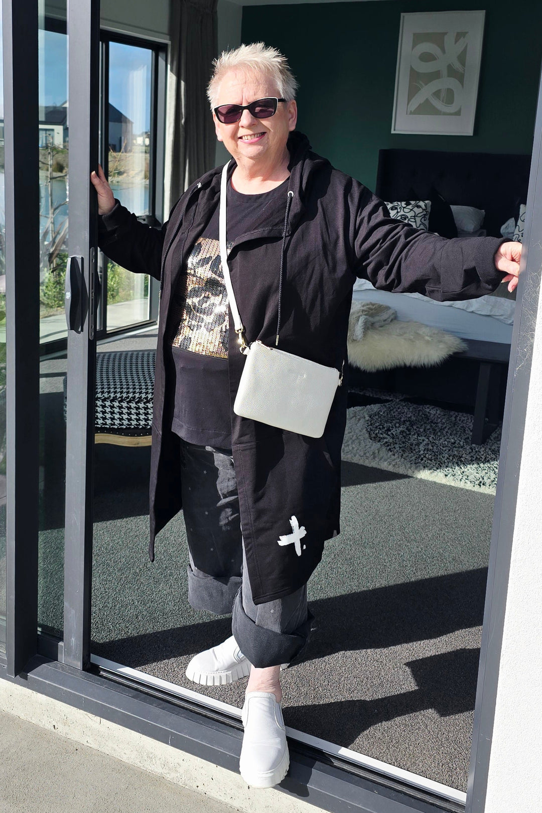 A woman wearing a black coat, sunglasses, and white sneakers stands in a modern bedroom doorway, holding the sliding glass door open with both hands. She is holding a small white shoulder bag.