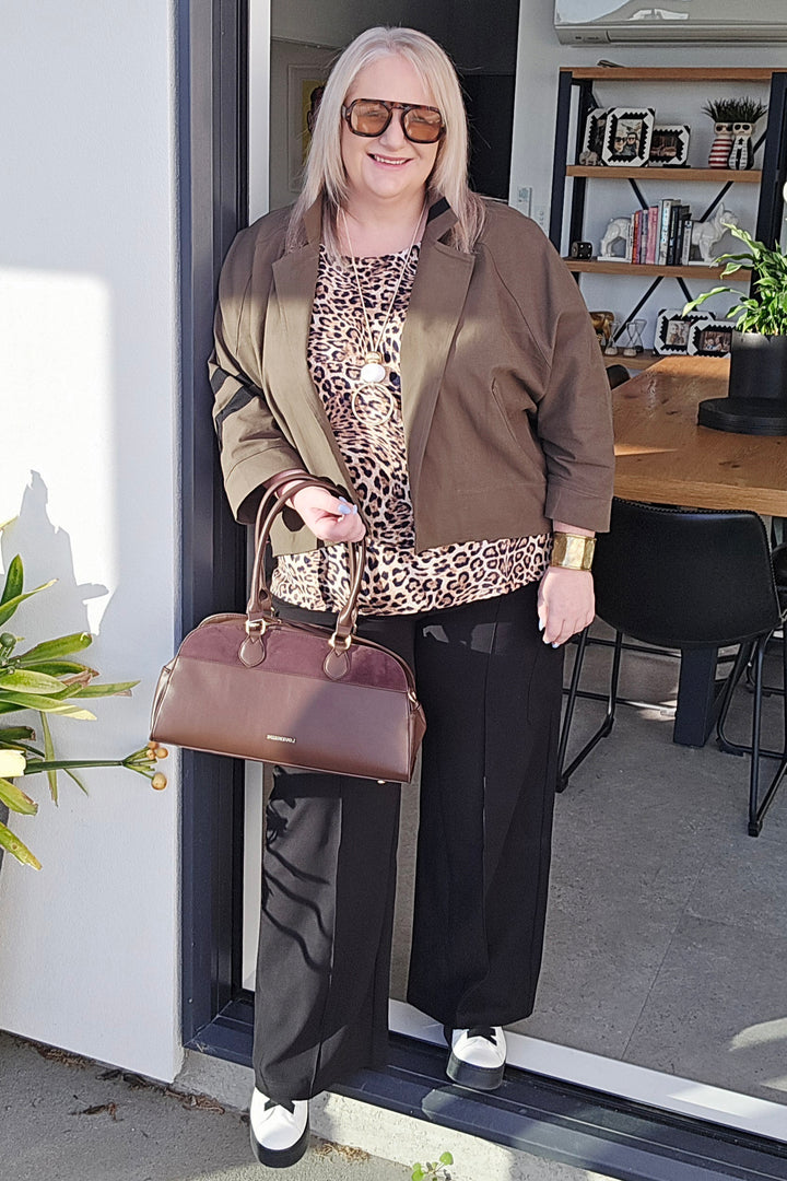 Woman holding a brown handbag, wearing a leopard print top and brown jacket, standing in front of a door.