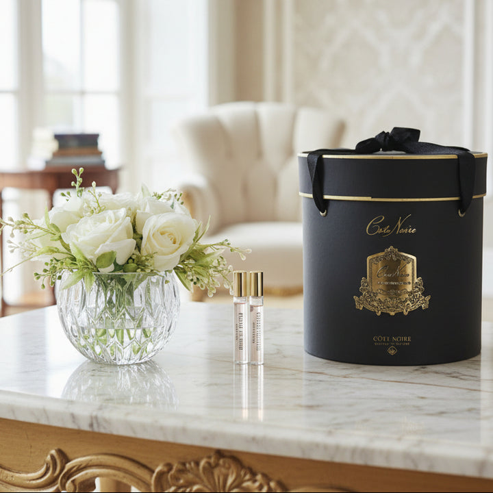 Cote Noire black round gift box with gold accents, a vase of flowers, and two bottles in a luxury room.