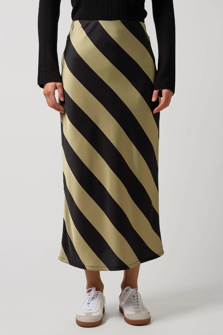 A model wearing a midi-length skirt with bold diagonal stripes in black and a warm beige/khaki color, styled with a black ribbed top and white sneakers against a light gray studio background.