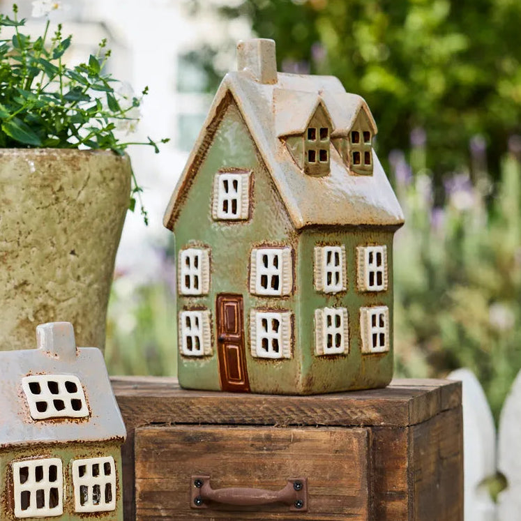 A decorative ceramic house-shaped figurine with a light green body, cream-colored textured trim, and a brown door, displayed on top of a rustic wooden chest outdoors.