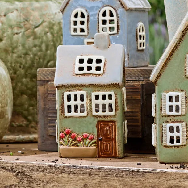 Three small ceramic house-shaped garden ornaments in pastel blue, sage green, and cream, displayed on a rustic wooden surface with other decorative vases in the background.