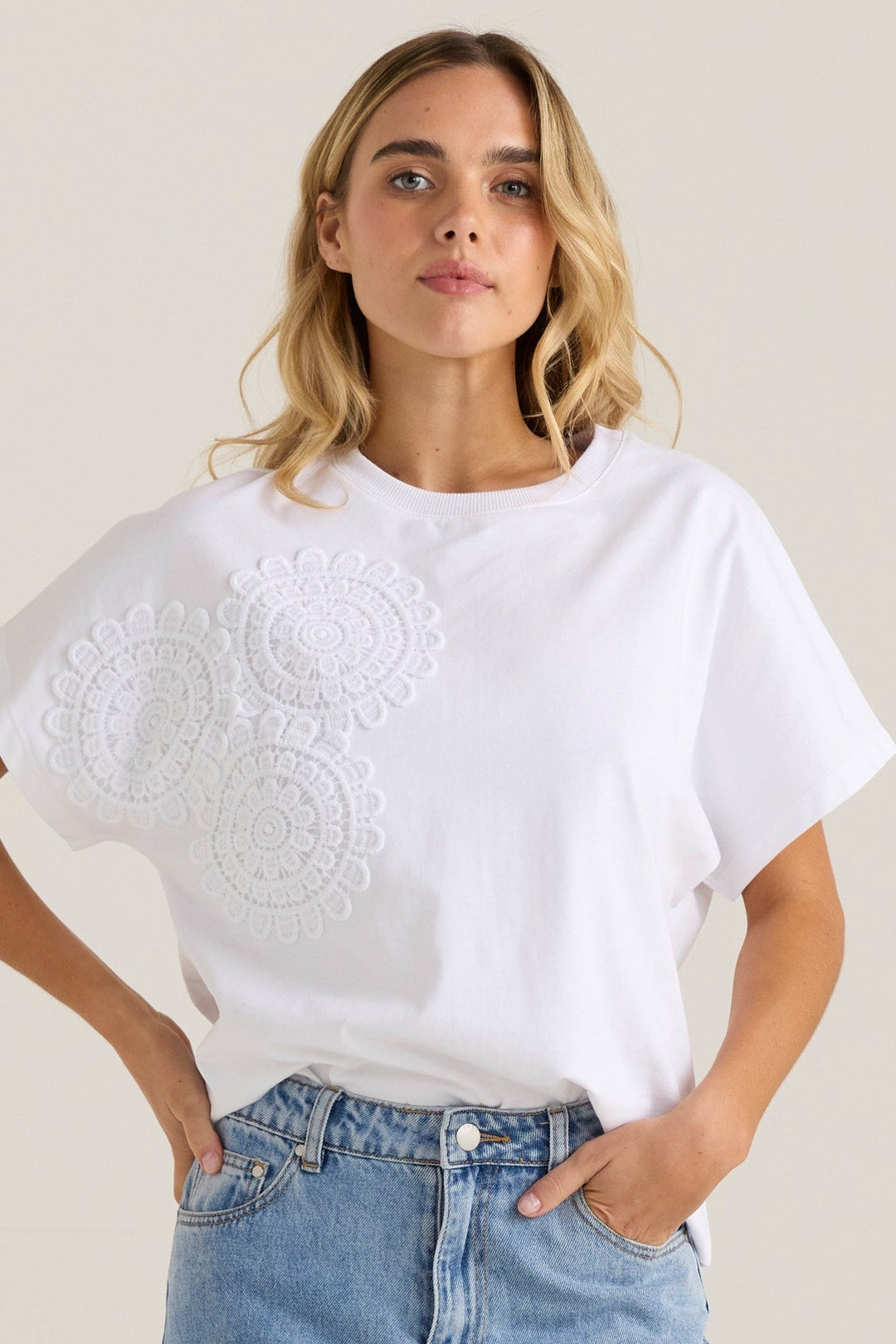 Woman wearing a white sass t-shirt with crochet details and blue jeans on a beige background