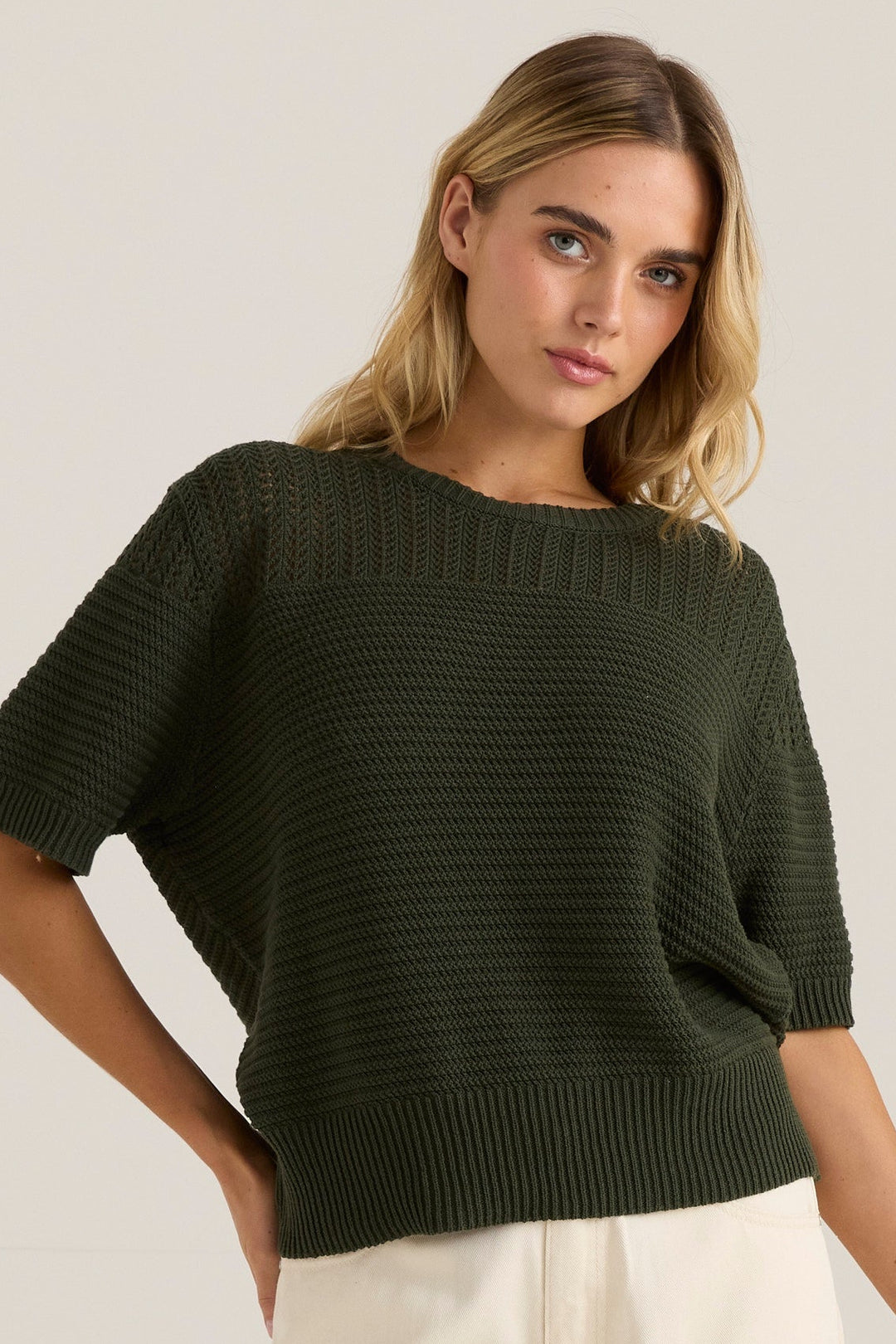 Woman wearing a dark green sass knit top and white pants against a plain background